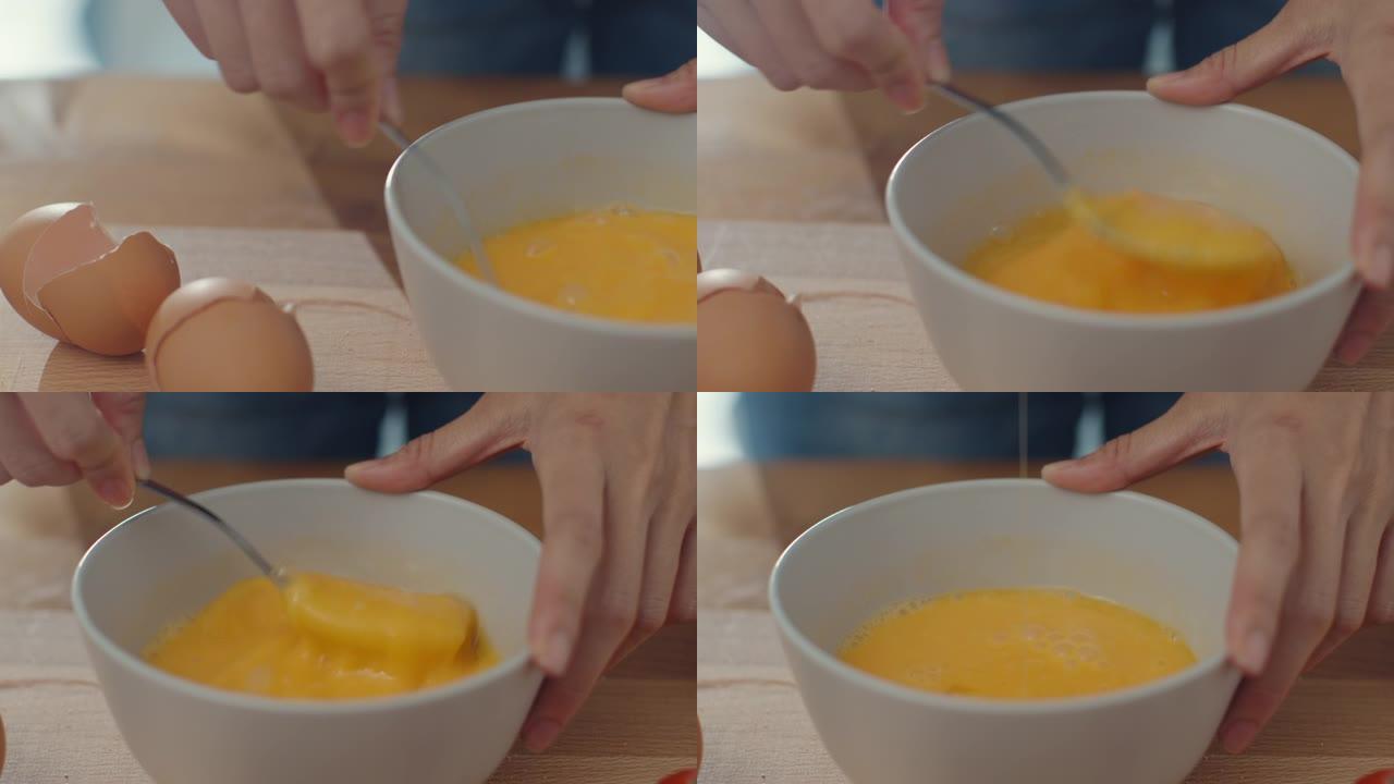 年轻的亚洲女厨师的手将鸡蛋搅拌到陶瓷碗中，在家里的厨房桌子上的木板上用蔬菜烹饪煎蛋。