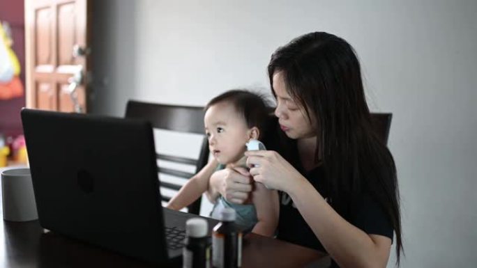 远程医疗亚洲华人母亲用体温计检查她的男婴的体温，同时用笔记本电脑在线咨询她的儿科医生