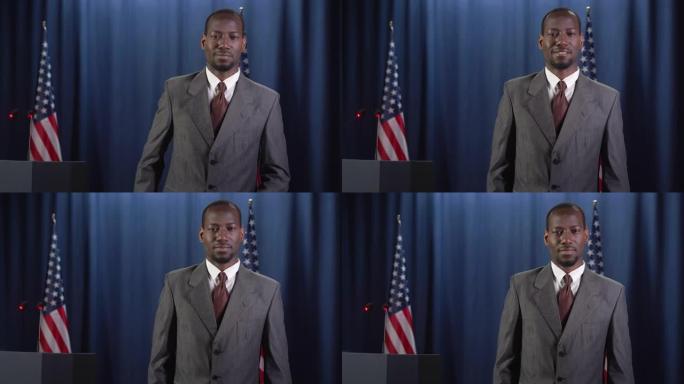 年轻的非洲裔美国政治家身穿灰色西装，面带微笑，在蓝色的美国国旗背景下对着镜头摆姿势