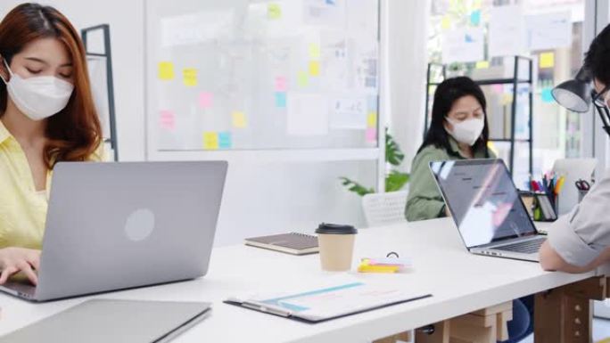 亚洲商人企业家戴着医用口罩，在新的正常情况下保持社交距离，以预防病毒，同时在办公室工作时使用笔记本电