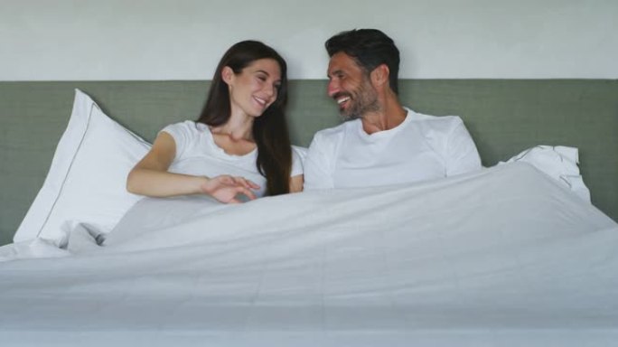 穿着睡衣的年轻新婚夫妇的真实照片是在卧室的床上一起休息时说话和笑的乐趣。早上刚醒来。