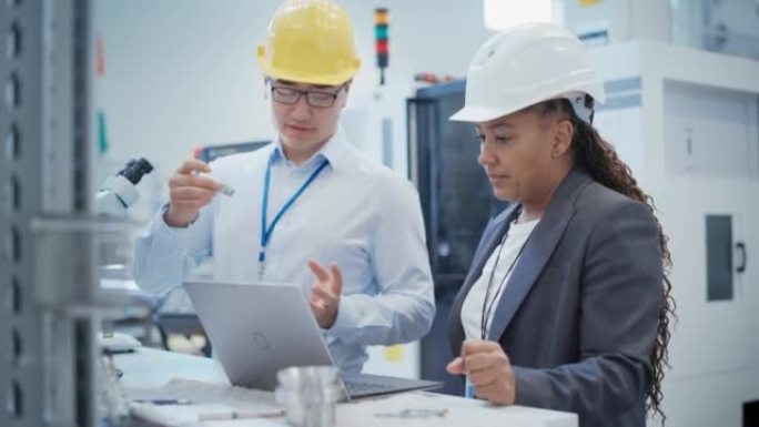 亚洲技术人员和非裔美国工程师戴着安全帽，在工厂使用笔记本电脑。讨论工业机器零件，将零件与草拟的蓝图进