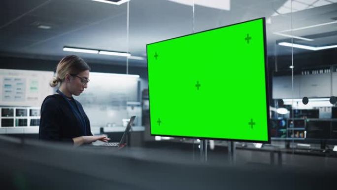 美丽的首席软件工程师独自站在会议室里，使用笔记本电脑，看着带有绿屏模拟显示的电视，其中包含开发部门创