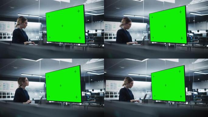 美丽的首席软件工程师独自站在会议室里，使用笔记本电脑，看着带有绿屏模拟显示的电视，其中包含开发部门创