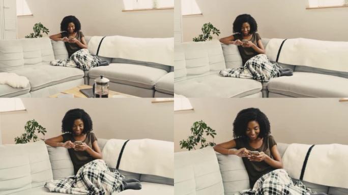 一名年轻的非洲裔美国妇女坐在家里的沙发上使用手机。一位美丽的混血女性，非洲裔在客厅的沙发上放松时上网