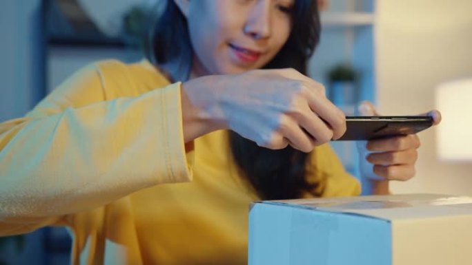 年轻的亚洲女性使用智能手机在包裹产品上拍摄条形码照片，以便晚上在家庭办公室运送给客户。
