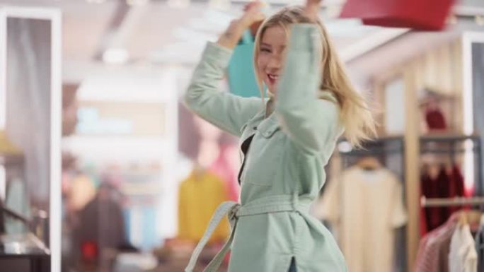 一个快乐的女性带着购物袋在服装店微笑、跳舞和旋转的肖像。年轻的成年金发白人女性顾客在百货商店购买时装