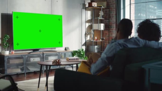 黑人夫妇在他们时尚的阁楼公寓里坐着看绿屏模拟显示的电视。周末娱乐概念。从后面开枪