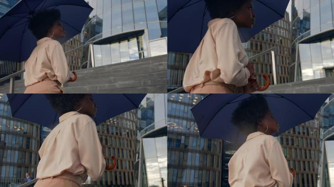 一个穿着米色服装的年轻迷人的黑皮肤卷发女人走上楼梯，在下雨天用两只手拿着深蓝色的雨伞