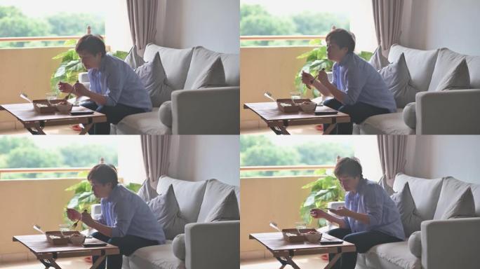 亚洲华裔妇女在客厅再生纸容器中使用智能手机阅读信息，一边享受外卖马来西亚食物nasi kerabu，