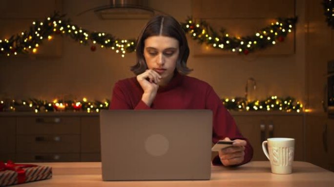 一位穿着红色毛衣的迷人年轻女子在网上商店的笔记本电脑上输入她的卡号，坐在厨房的桌子旁