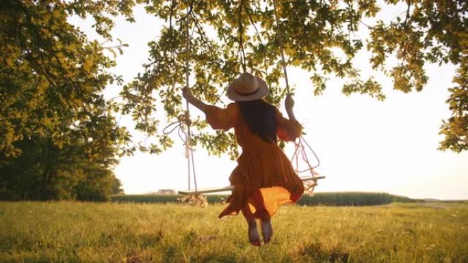 特写，手持镜头，一名穿着红色连衣裙的年轻成年女性摇摆，坐在悬挂在阳光充足的草地上的大树枝上的绳索秋千