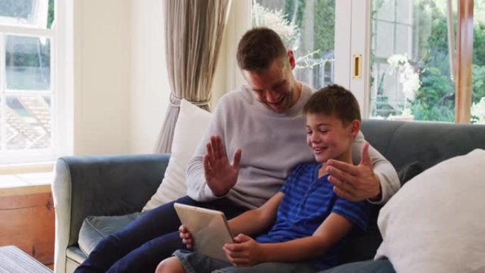 白人男子和儿子在坐在ho的沙发上的数字平板电脑上进行视频通话时微笑着