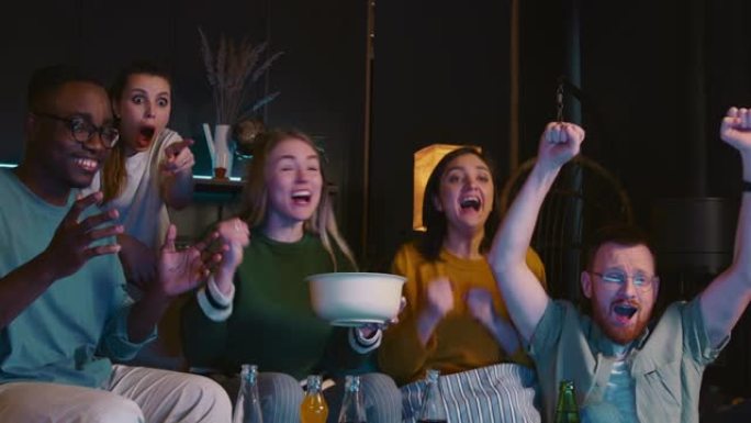 一群年轻快乐的多民族体育迷在家里用零食和饮料在电视上观看比赛，庆祝胜利和胜利