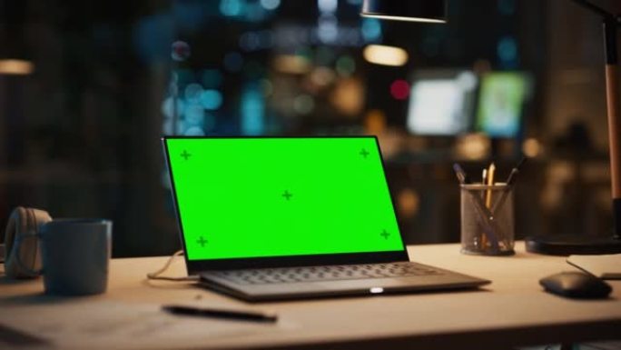 笔记本电脑站在桌子上，配有绿屏Chromakey模拟显示器。晚上创意办公室配有电脑、咖啡杯、耳机和笔