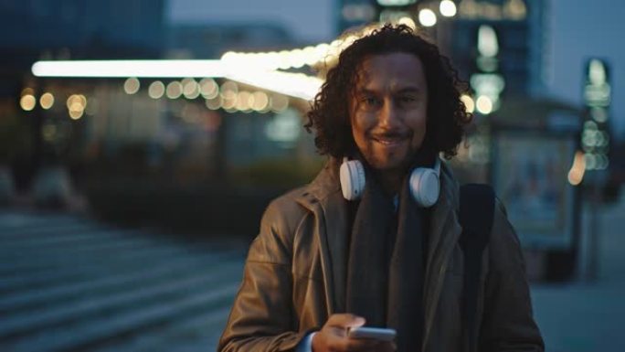 戴着无线耳机的SLO MO西班牙裔男子在城市的人行道上摆姿势