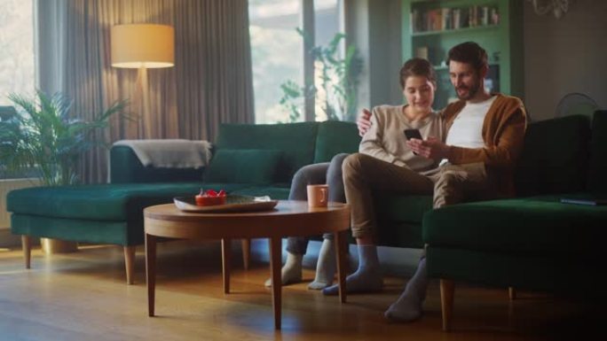 夫妇坐在舒适公寓的沙发上时，使用智能手机设备。男女朋友聊天，在互联网上进行电子购物，观看有趣的视频，