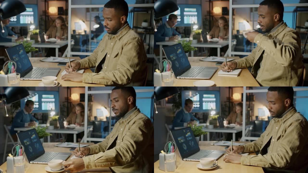 非裔美国商人看着笔记本电脑屏幕做笔记，晚上在办公室喝咖啡