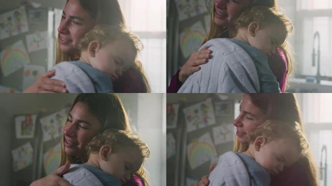 年轻的快乐和平母亲的电影镜头正在拥抱和唱歌摇篮曲她的蹒跚学步的男婴，同时睡在家里的厨房里。甜梦、童年