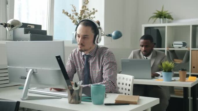 快乐的员工使用电脑打字，然后在办公室通过耳机跳舞欣赏音乐