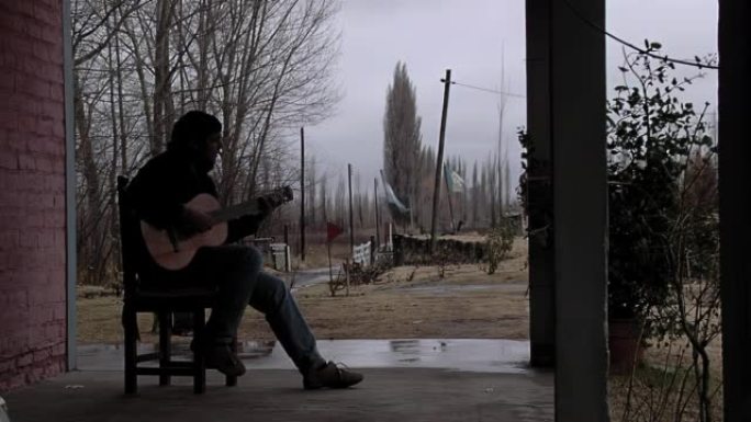 一名男子在他家的门廊里在户外弹吉他，背景是阿根廷和内乌肯省的旗帜。