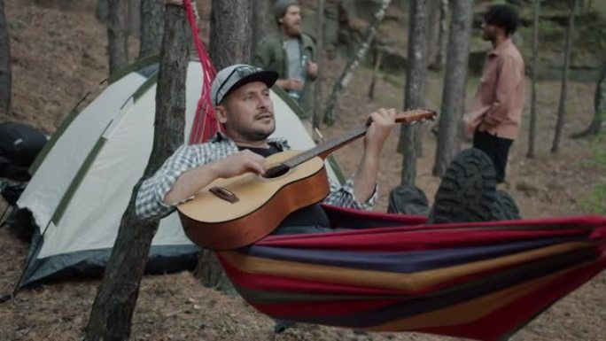 快乐的家伙在营地的吊床上弹吉他放松，徒步旅行者在背景中行走