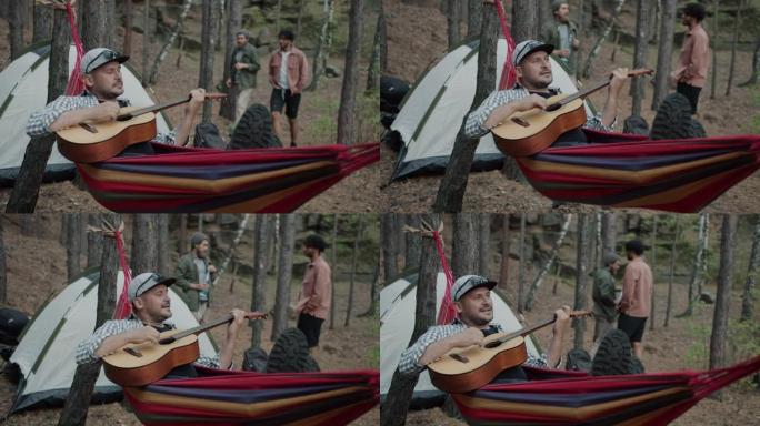 快乐的家伙在营地的吊床上弹吉他放松，徒步旅行者在背景中行走