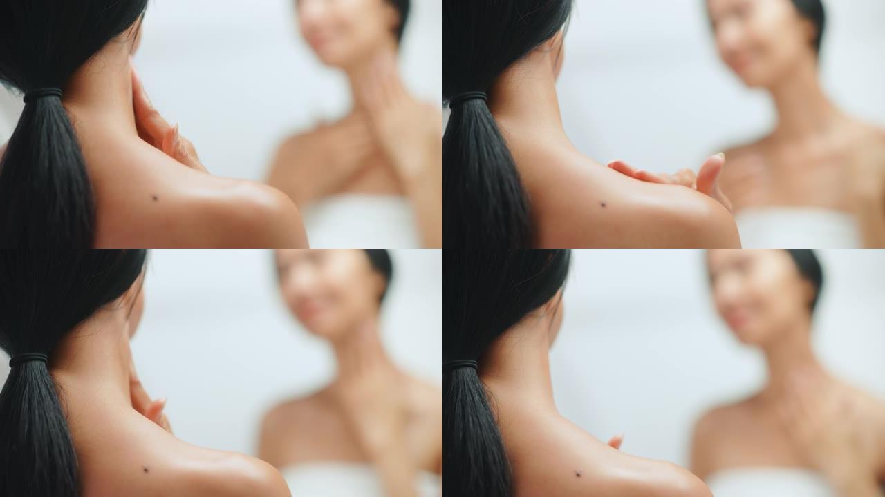 美丽的亚洲女人的肖像用性感的手势轻轻地涂抹润肤霜，看着浴室的镜子。年轻女性用天然化妆品护肤产品使皮肤