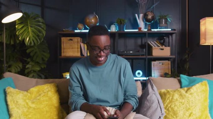 年轻快乐的微笑z世代黑人男学生点头，在家里的沙发上使用智能手机应用程序随着音乐跳舞。