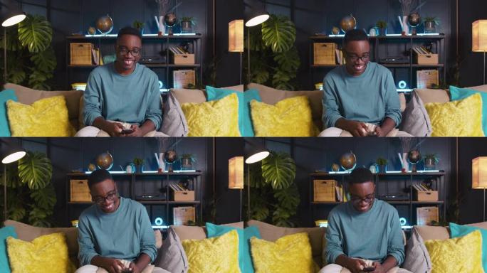 年轻快乐的微笑z世代黑人男学生点头，在家里的沙发上使用智能手机应用程序随着音乐跳舞。