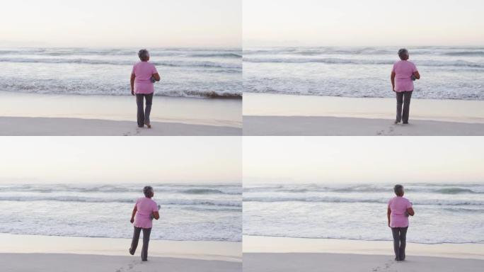 非裔美国高级妇女站在沙滩上时拿着瑜伽垫看着海浪