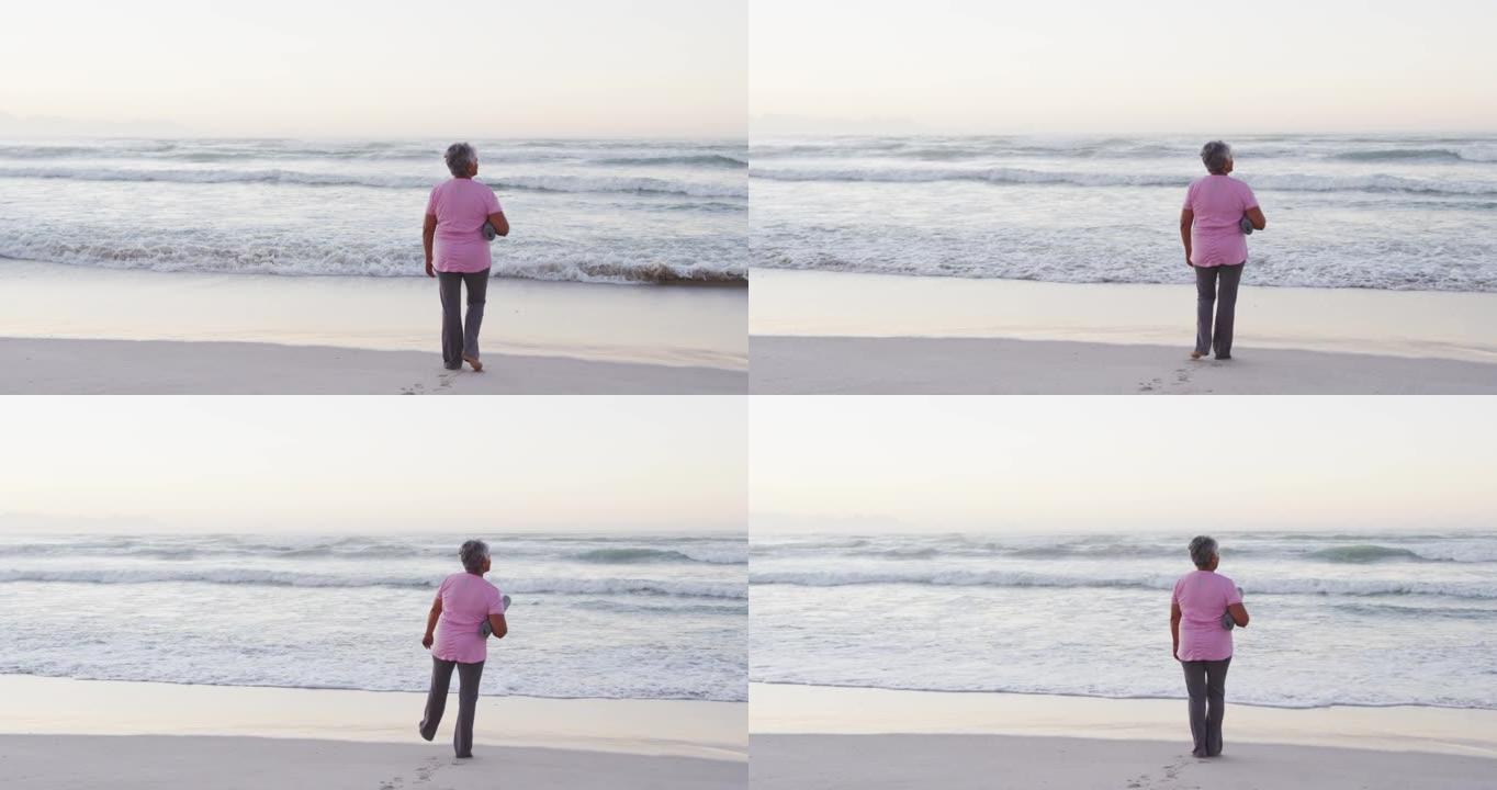 非裔美国高级妇女站在沙滩上时拿着瑜伽垫看着海浪
