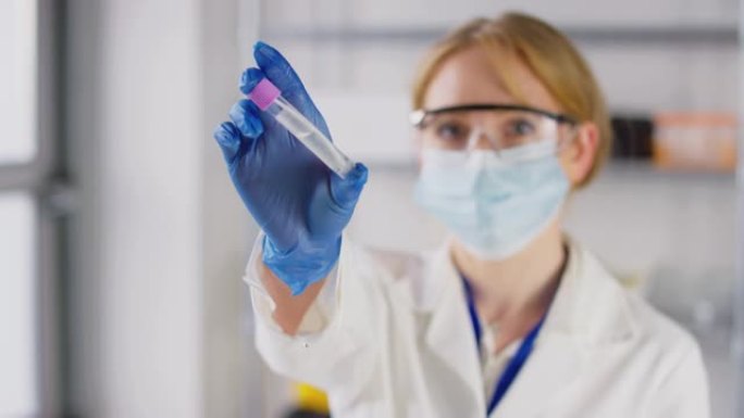 拉焦点肖像的女实验室研究人员戴着安全眼镜和面罩，拿着含有PCR拭子测试的试管，以慢镜头拍摄