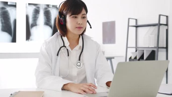 年轻的亚洲女医生，身穿白色医疗制服，带听诊器，使用电脑笔记本电脑，在诊所或医院的办公桌上与病人进行视
