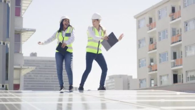 两个美丽的快乐自信的多民族年轻女工程师跳舞，拿着数字平板电脑和蓝图，计划，示意图，同时在建筑工地安装