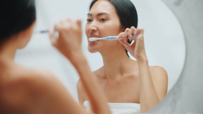 美丽的亚洲女人的肖像用牙刷清洁她的天然洁白的牙齿，微笑在浴室的镜子里看着相机。做早上例行公事的快乐女