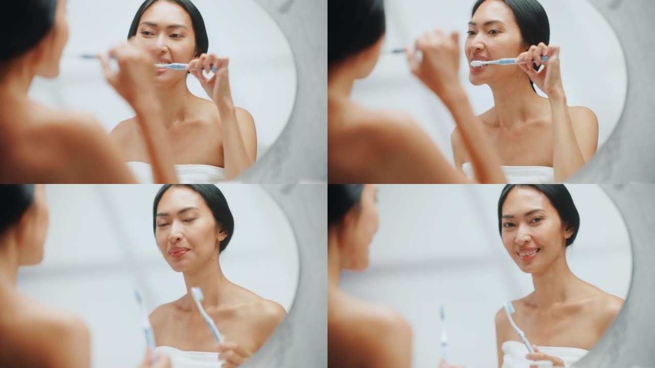 美丽的亚洲女人的肖像用牙刷清洁她的天然洁白的牙齿，微笑在浴室的镜子里看着相机。做早上例行公事的快乐女