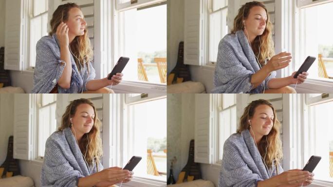 快乐的白人妇女戴着耳机坐在阳光明媚的小屋客厅使用智能手机