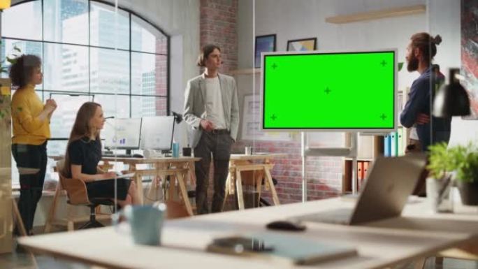 时尚的年轻项目经理在创意机构会议室的团队会议上讲话。同事在绿屏模拟色度键显示器上显示营销计划演示。