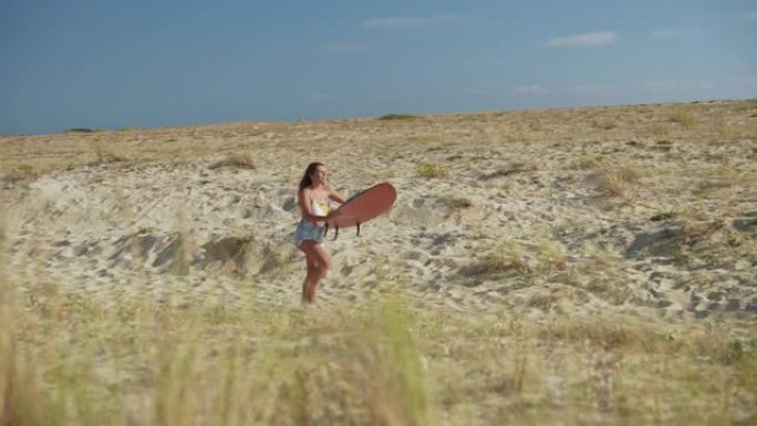 女人用冲浪板在手臂下行走，在沙丘到海洋之间