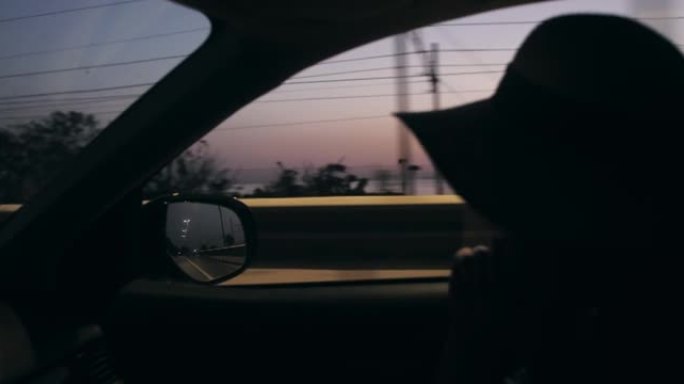 在日出海路慢动作驾驶的汽车乘客座位上戴着夏日帽子的美女特写剪影拍摄。