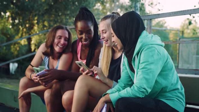 一群年轻的多种族女性青少年坐在公园里，在智能手机上观看有趣的视频。一群朋友在户外一起玩乐，同时保持联
