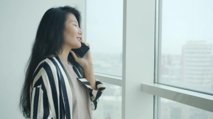 开朗的亚洲女人在手机上聊天，在公寓的大窗户附近微笑