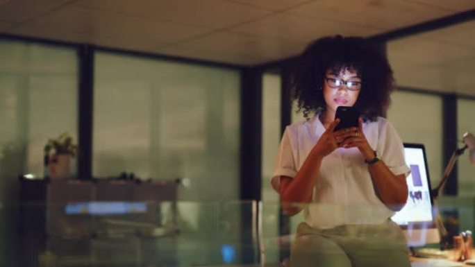 年轻的女商人在办公室工作到很晚的时候在电话上在线打字。一位公司女性专业人士独自在工作中加班时在互联网