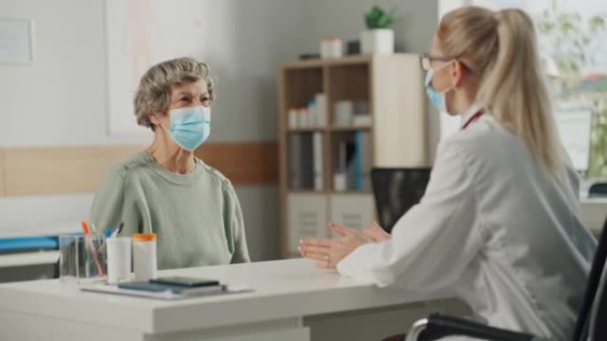 女家庭医生在健康诊所咨询期间对一位资深妇女讲话。两人都戴着口罩。穿着白大褂的医生在医院办公室给老年病