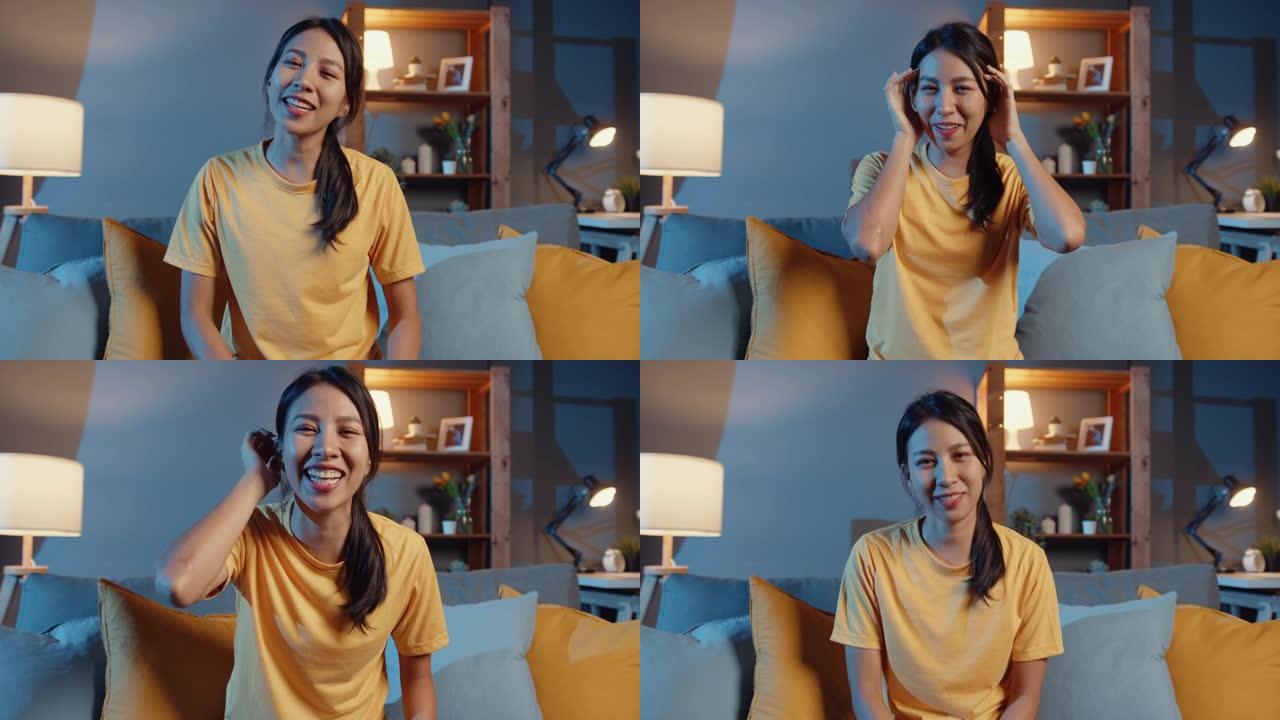 快乐的年轻自由亚洲女性看着相机微笑，晚上在家里的客厅在线视频通话与朋友交谈。