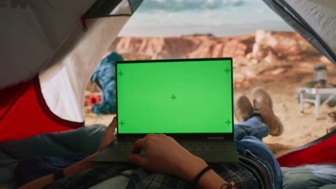 游客使用带有绿屏占位符的笔记本电脑的POV镜头。旅行者在落基山顶上的帐篷里休息。冒险的徒步旅行者生活