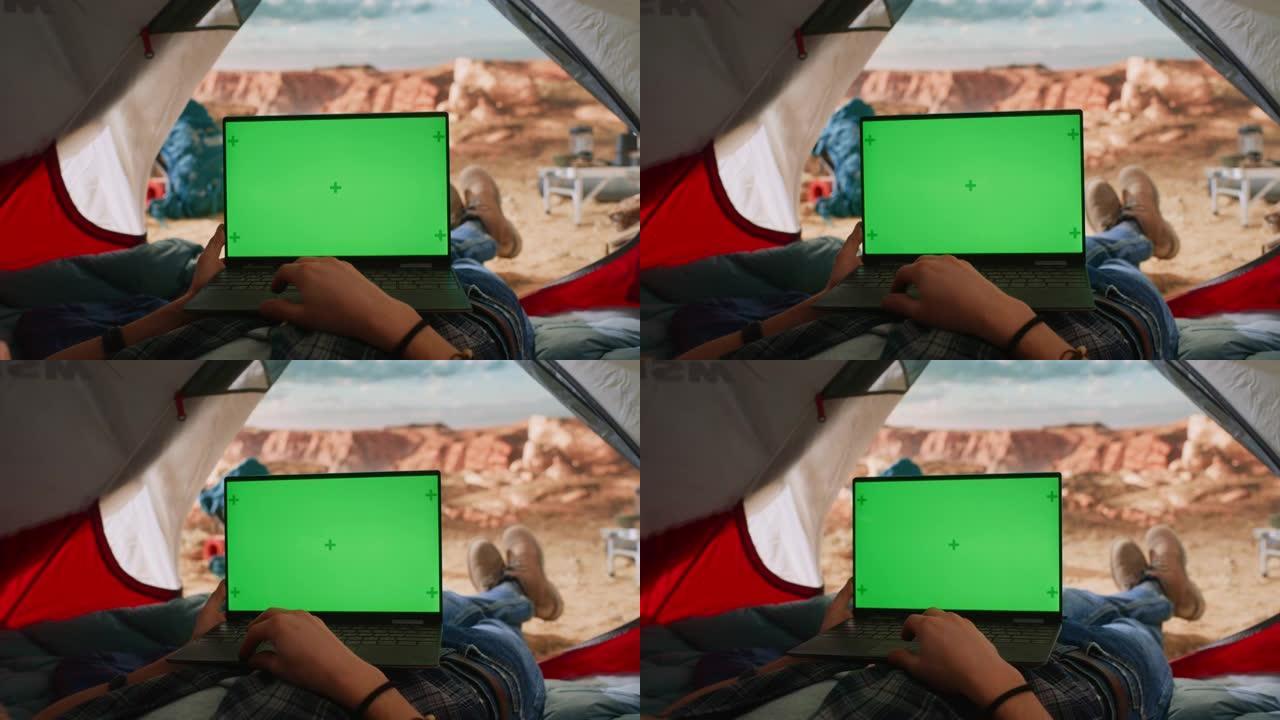 游客使用带有绿屏占位符的笔记本电脑的POV镜头。旅行者在落基山顶上的帐篷里休息。冒险的徒步旅行者生活