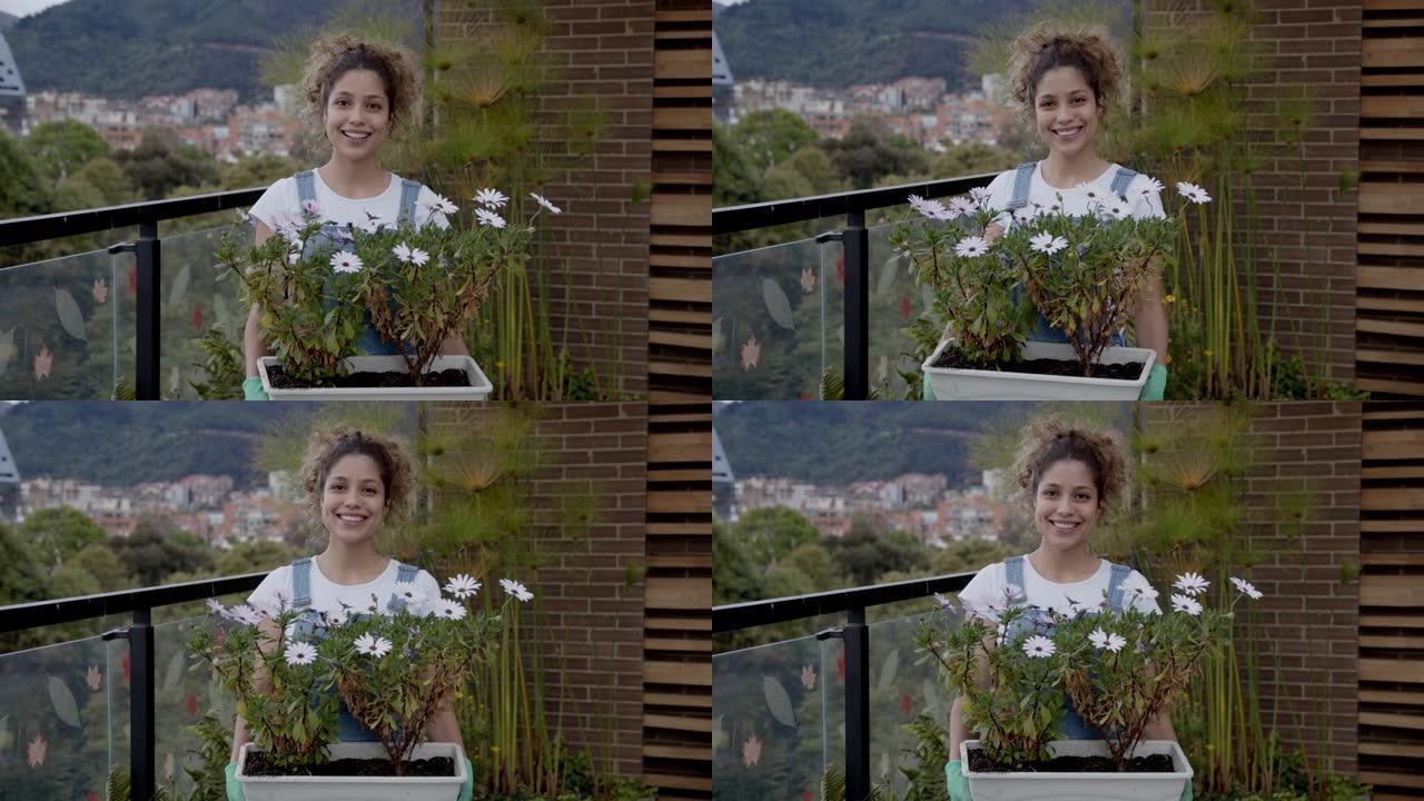 快乐的年轻女子园艺在她的阳台看相机微笑，而拿着一个花盆