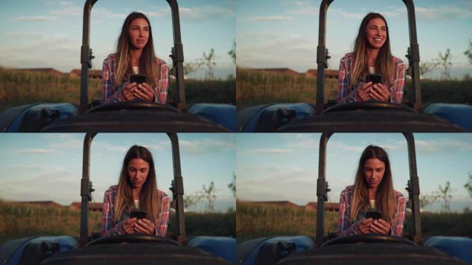 日落时坐在拖拉机上，微笑着笑着使用智能手机的女人的肖像。女性农民保持联系并使用互联网改善业务